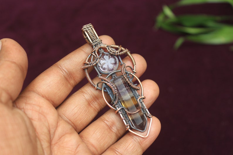 May include: A wire-wrapped pendant featuring a brown and clear crystal with a smaller, clear crystal with a flower design. The pendant is made with copper and silver wire.