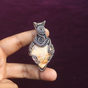 May include: A wire-wrapped pendant featuring a light brown stone with a unique pattern. The pendant is made with copper wire and has a delicate, intricate design.