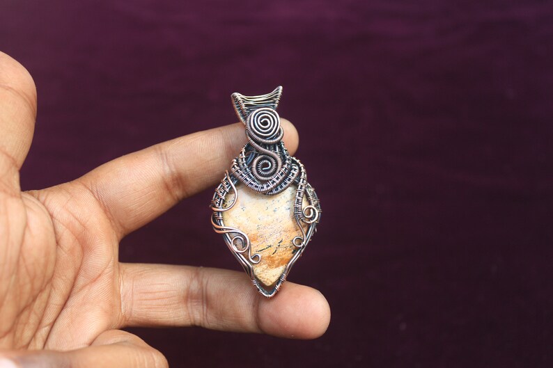 May include: A wire-wrapped pendant featuring a light brown stone with a unique pattern. The pendant is made with copper wire and has a delicate, intricate design.
