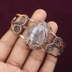 May include: A handcrafted cuff bracelet featuring a large, oval-shaped, clear gemstone with black inclusions. The bracelet has a copper and silver-toned metal design with intricate swirls and decorative accents. The bracelet is approximately 2 inches wide.