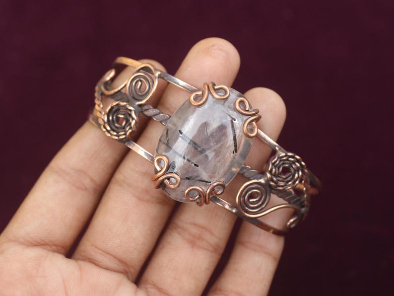 May include: A handcrafted cuff bracelet featuring a large, oval-shaped, clear gemstone with black inclusions. The bracelet has a copper and silver-toned metal design with intricate swirls and decorative accents. The bracelet is approximately 2 inches wide.