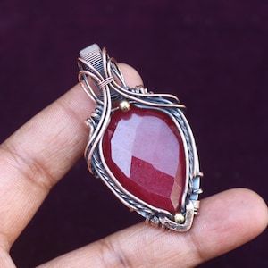 May include: A handcrafted pendant featuring a faceted, teardrop-shaped, deep red gemstone. The stone is encased in a wire-wrapped setting with silver and copper-coloured metal accents. Small gold-coloured beads add detail to the design.