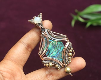 Könnte beinhalten: Ein Anhänger aus Kupfer- und Silberdraht, der mit einer grün-blauen Abalone-Muschel mit einem weißen Opal-Akzent umwickelt ist.