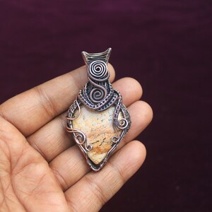 May include: A copper wire wrapped pendant featuring a light brown and white agate stone. The pendant is in the shape of a teardrop and has a detailed wire design.