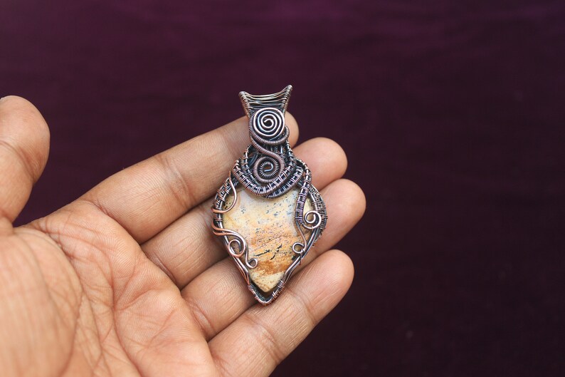 May include: A copper wire wrapped pendant featuring a light brown and white agate stone. The pendant is in the shape of a teardrop and has a detailed wire design.