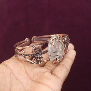 May include: Copper-toned cuff bracelet featuring a large, clear gemstone with black inclusions. The bracelet has intricate wirework and decorative swirls. The gemstone is oval-shaped and the bracelet is held in a hand.