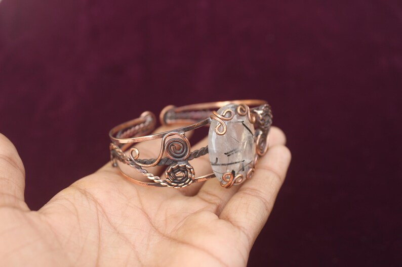 May include: Copper-toned cuff bracelet featuring a large, clear gemstone with black inclusions. The bracelet has intricate wirework and decorative swirls. The gemstone is oval-shaped and the bracelet is held in a hand.
