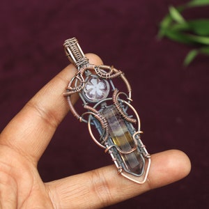 May include: A copper wire wrapped pendant featuring a clear quartz crystal and a brown and yellow fluorite crystal. The pendant is held by a hand.