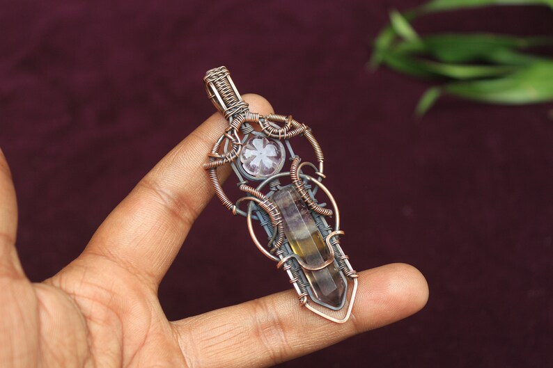May include: A copper wire wrapped pendant featuring a clear quartz crystal and a brown and yellow fluorite crystal. The pendant is held by a hand.