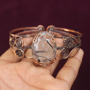 May include: Copper-toned cuff bracelet featuring an oval, clear gemstone with black inclusions. The bracelet has intricate wirework and spiral designs, with a dark background.