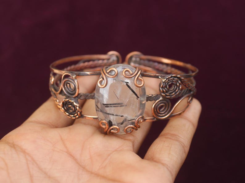 May include: Copper-toned cuff bracelet featuring an oval, clear gemstone with black inclusions. The bracelet has intricate wirework and spiral designs, with a dark background.