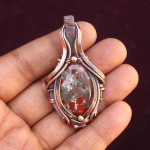 May include: A copper wire wrapped pendant featuring a red, white, and gray stone with a speckled pattern. The pendant has a gold accent at the top.