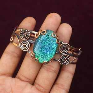 May include: A handcrafted cuff bracelet with a central oval-shaped dichroic glass stone in shades of teal and blue. The metalwork features intricate swirls and rosettes in silver and copper tones. The bracelet is approximately 2 inches wide.