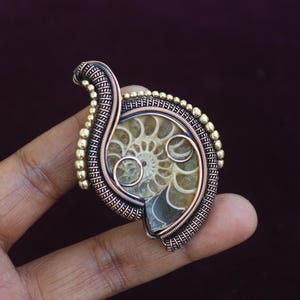 May include: A handcrafted pendant featuring a fossilized ammonite encased in copper wire. The pendant has a spiral design with a bronze and gold-colored wire frame. The fossil is a light brown color and is held in place by the wire.