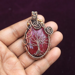 May include: A copper wire wrapped pendant featuring a red jasper stone with a tree of life design. The pendant has gold accents and is held in a hand.