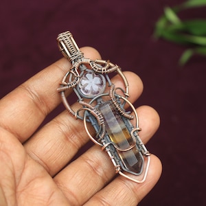 May include: A wire-wrapped pendant featuring a brown and clear crystal with a smaller, clear crystal with a flower design. The pendant is made with copper and silver wire.