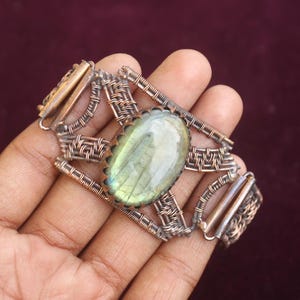 May include: A handcrafted copper bracelet featuring a large, oval-shaped labradorite stone. The stone is a pale green color with a scalloped edge. The bracelet has an intricate wire-wrapped design.