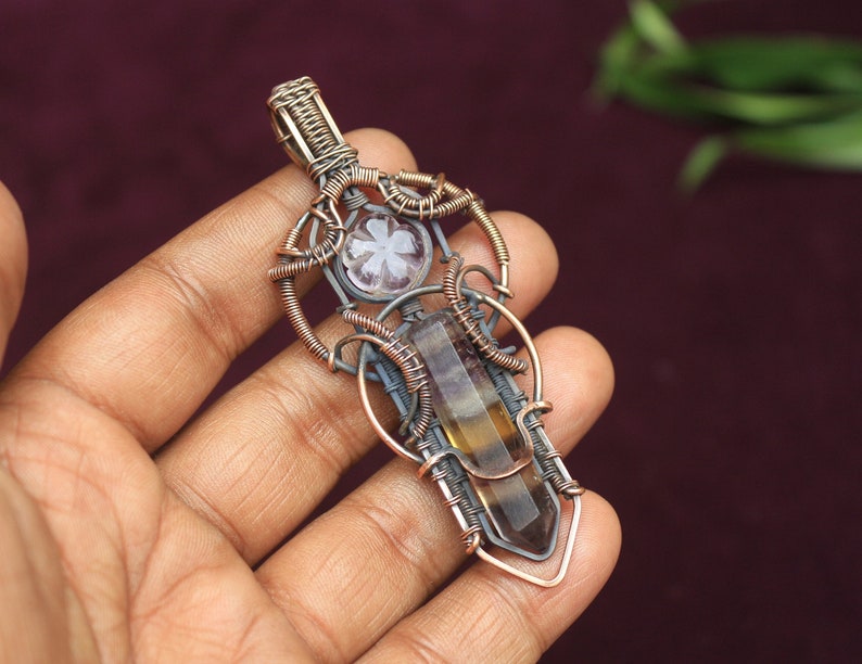 May include: A wire-wrapped pendant featuring a clear and brown fluorite crystal and a white flower-shaped stone. The pendant is made with copper wire and has a silver-toned finish.