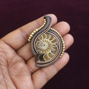 May include: A handcrafted pendant featuring a fossilized ammonite encased in a wire-wrapped setting. The pendant showcases a spiral pattern in shades of brown and tan, with gold-colored accents. The wirework is intricate, creating a unique, artistic piece.