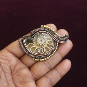May include: A handcrafted pendant featuring a fossilized ammonite encased in copper wire. The pendant has a spiral design with a bronze and gold color scheme. The ammonite fossil is visible in the center.