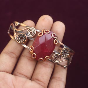 Pode incluir: Pulseira de cobre artesanal com uma grande pedra preciosa vermelha facetada no centro. A pulseira apresenta intrincados trabalhos de arame com desenhos em espiral e florais. O metal tem uma mistura de tons de cobre e prata.
