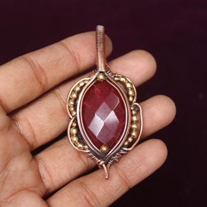Puede incluir: Un colgante artesanal con una piedra preciosa facetada de color rojo intenso. La piedra está engastada en un marco de alambre de cobre y plata, acentuado con pequeñas cuentas doradas. El colgante tiene un lazo para una cadena.