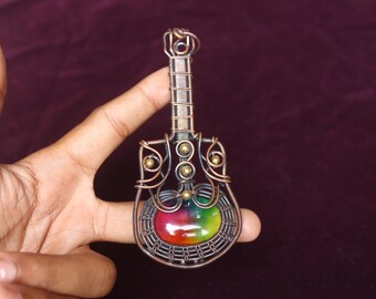Puede incluir: Colgante de guitarra hecho a mano con alambre de cobre, con una piedra de colores del arcoíris en el centro. El colgante tiene un diseño detallado de alambre enrollado con acentos dorados, creando una pieza de joyería única y artística.