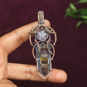 May include: A copper wire wrapped pendant featuring a fluorite crystal and a smaller clear crystal. The pendant is in the shape of a sword with intricate wirework.
