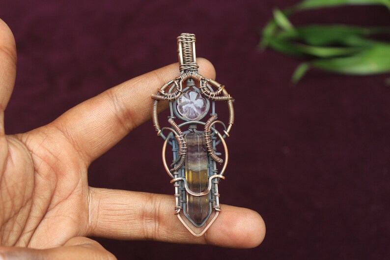 May include: A copper wire wrapped pendant featuring a fluorite crystal and a smaller clear crystal. The pendant is in the shape of a sword with intricate wirework.