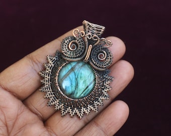 Puede incluir: Colgante artesanal con una piedra preciosa redonda, iridiscente azul-verdosa, intrincadamente alambrada en cobre. El colgante tiene un diseño de sol con detalles en espiral, creando una pieza de joyería artística única.