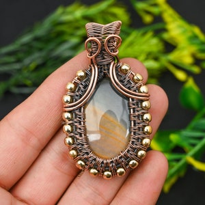 May include: A wire-wrapped pendant featuring a light brown agate stone with gold and copper wire accents. The pendant is oval-shaped and has a delicate, intricate design.