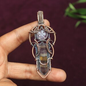 May include: A copper wire wrapped pendant featuring a fluorite crystal and a smaller clear crystal. The pendant is in the shape of a sword with intricate wirework.