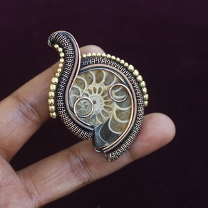 May include: A handcrafted pendant featuring a fossilized ammonite encased in intricate wire wrapping. The pendant showcases a spiral pattern in shades of beige and brown, accented by copper and gold-toned metal. A unique piece of jewelry.