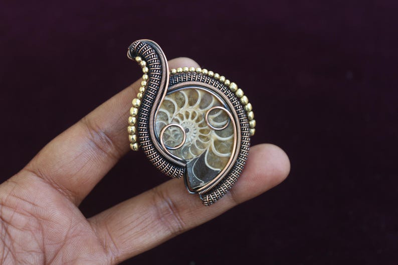 May include: A handcrafted pendant featuring a fossilized ammonite encased in intricate wire wrapping. The pendant showcases a spiral pattern in shades of beige and brown, accented by copper and gold-toned metal. A unique piece of jewelry.