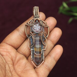 May include: A wire-wrapped pendant featuring a multi-colored fluorite crystal and a smaller, clear crystal. The pendant is made with copper and silver wire and has a geometric design.