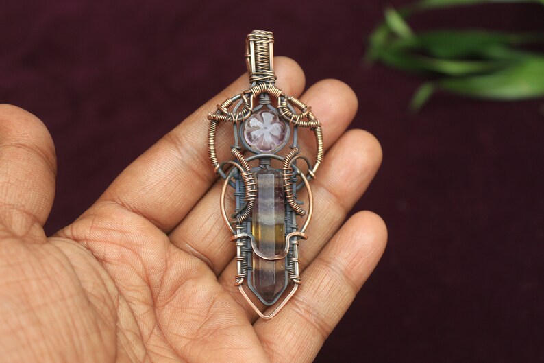 May include: A wire-wrapped pendant featuring a multi-colored fluorite crystal and a smaller, clear crystal. The pendant is made with copper and silver wire and has a geometric design.
