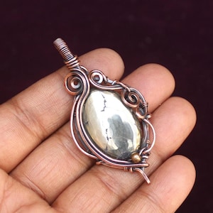 May include: A handcrafted copper pendant with a polished, oval-shaped pyrite stone. The stone is encased in intricate copper wirework, featuring swirls and loops. The pendant is held in a hand against a dark background, showcasing the jewelry's details.