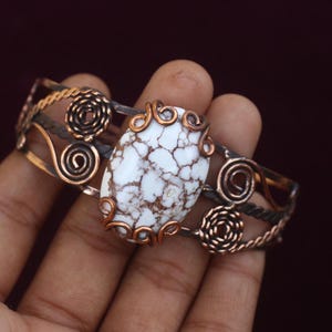 May include: A handcrafted copper bracelet featuring a large, oval white stone with brown veining. The bracelet has intricate wirework and spiral accents, showcasing a unique, artistic design. The metal has a warm, antique finish.