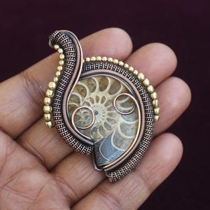 May include: A handcrafted pendant featuring a fossilized ammonite encased in copper wire. The pendant has a spiral design with a bronze and gold-colored wire wrap. The fossil is a light tan color with a dark brown background.