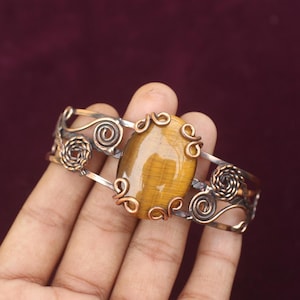 May include: A handcrafted cuff bracelet featuring a large, oval tiger's eye stone. The stone is set in a silver-toned metal with copper accents and decorative swirls. The bracelet is approximately 2 inches wide.