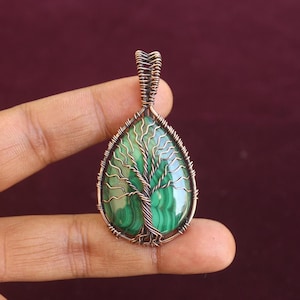 Puede incluir: Colgante de cobre hecho a mano con una piedra de malaquita en forma de lágrima y un diseño de árbol alambrado. El colgante está suspendido de una fianza de cobre, mostrando los intrincados detalles del árbol de la vida.