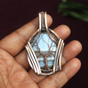 May include: A handcrafted copper pendant featuring a light blue stone with a wire-wrapped tree design. The pendant has a detailed, openwork frame and is held in a hand, showcasing the intricate craftsmanship and natural elements.