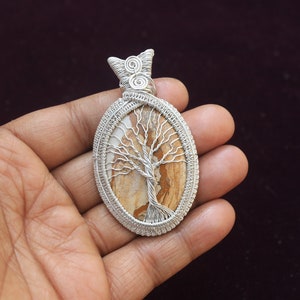 May include: A silver wire wrapped pendant featuring a brown and white tree of life design. The pendant is oval shaped and has a silver bail.