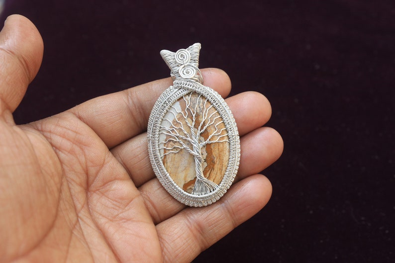 May include: A silver wire wrapped pendant featuring a brown and white tree of life design. The pendant is oval shaped and has a silver bail.