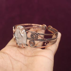 May include: Copper cuff bracelet featuring an oval, clear gemstone with black inclusions. The gemstone is set in a decorative copper frame with swirling accents. The bracelet has a detailed, openwork design with twisted and coiled copper elements.
