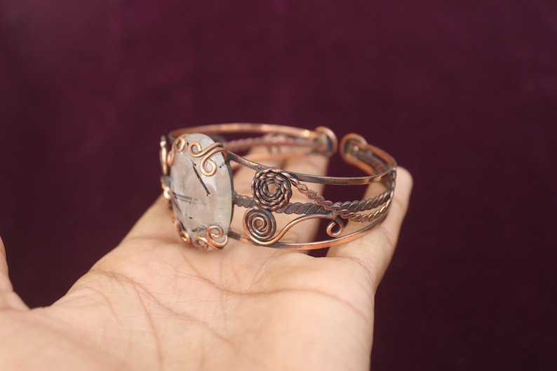 May include: Copper cuff bracelet featuring an oval, clear gemstone with black inclusions. The gemstone is set in a decorative copper frame with swirling accents. The bracelet has a detailed, openwork design with twisted and coiled copper elements.