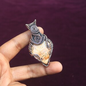 May include: A pendant featuring a light brown stone wrapped in copper wire with intricate designs. The wire is twisted and curled, creating a unique and eye-catching piece of jewelry.