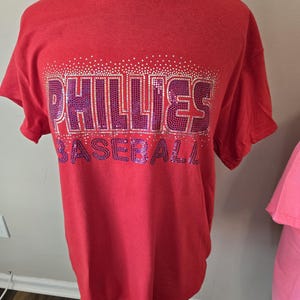 May include: Red t-shirt with a purple and white rhinestone design that says "Phillies Baseball".