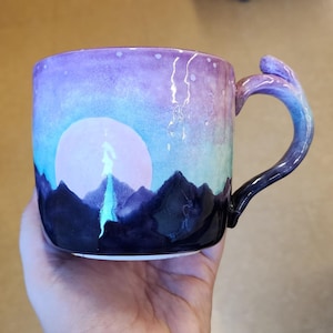 Puede incluir: Una taza de cerámica con un diseño de paisaje pintado a mano. La taza presenta un cielo degradado púrpura, azul y verde con una gran luna rosa pálido. Montañas azul oscuro en la base. El asa tiene una forma curva única.