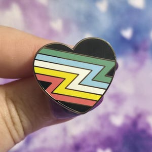Könnte beinhalten: Eine herzförmige Emaille-Anstecknadel mit schwarzem Hintergrund und einem horizontalen Zickzackmuster in den Farben Grün, Hellblau, Weiß, Gelb und Koralle. Die Anstecknadel hat einen dünnen Metallrand und eine glatte, glänzende Oberfläche.